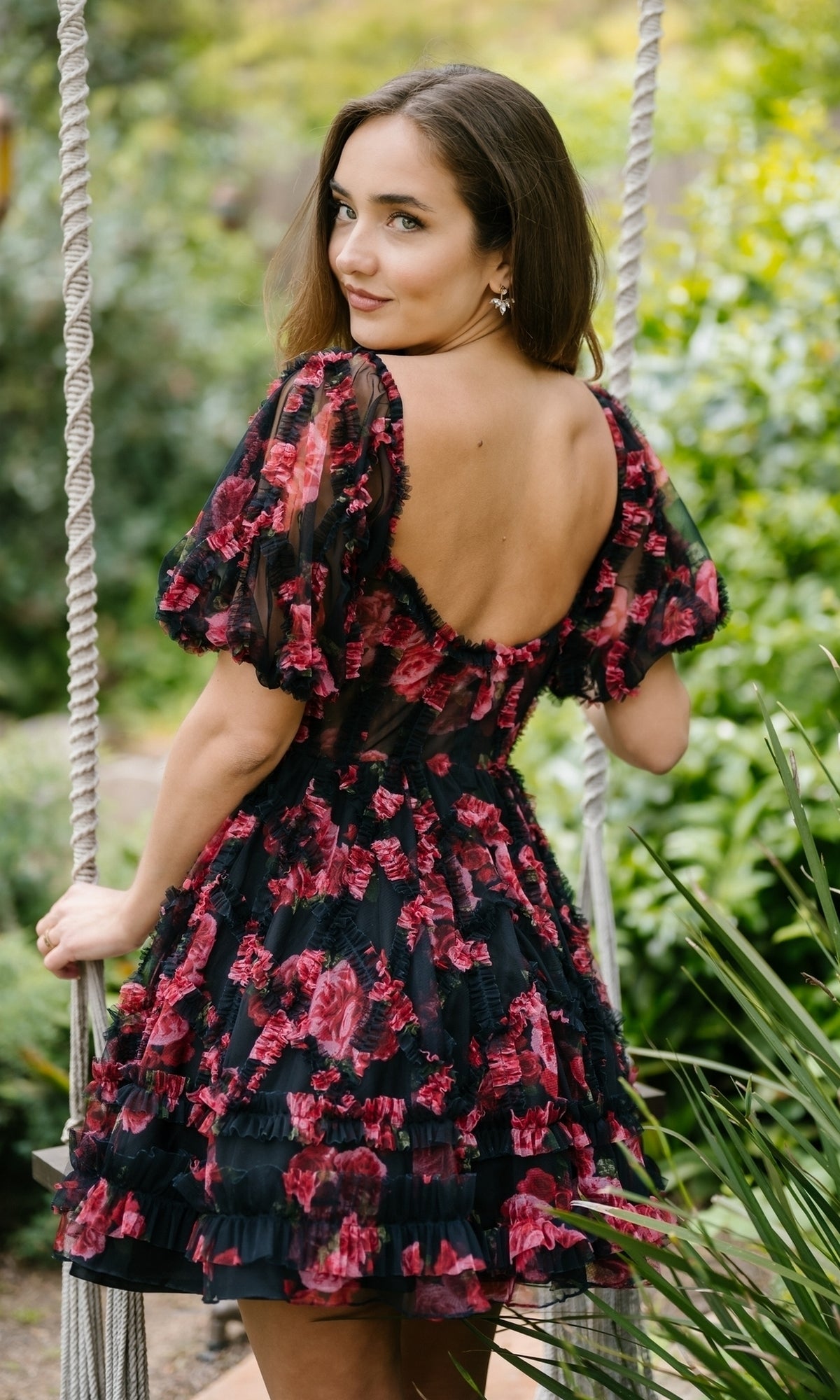 A woman wearing the Aleta 1304 Short Sleeve Short Print Prom Dress stands outdoors, holding rope swings and glancing over her shoulder in a garden setting.